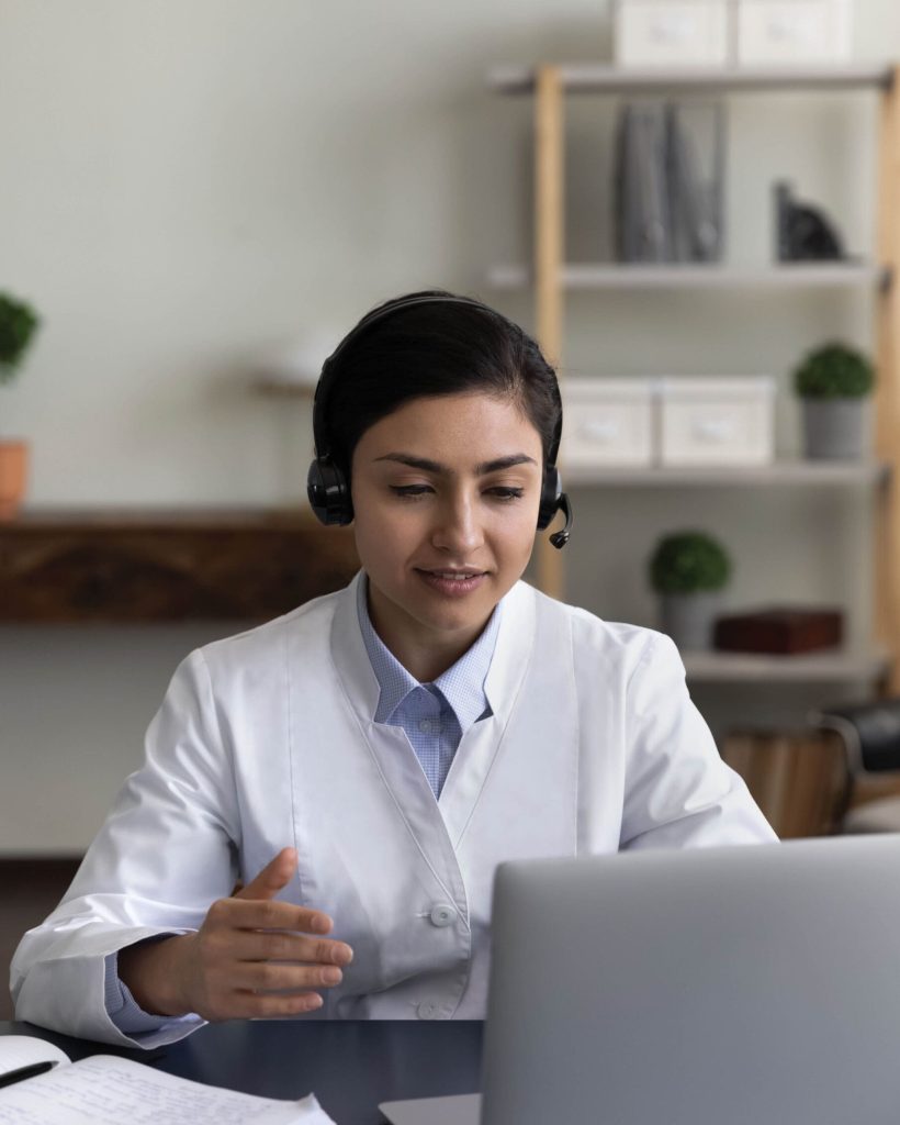 Vertical photo of young Indian woman doctor in headphones talking, using laptop in office, female physician general practitioner consulting patient online by video call, intern watching webinar