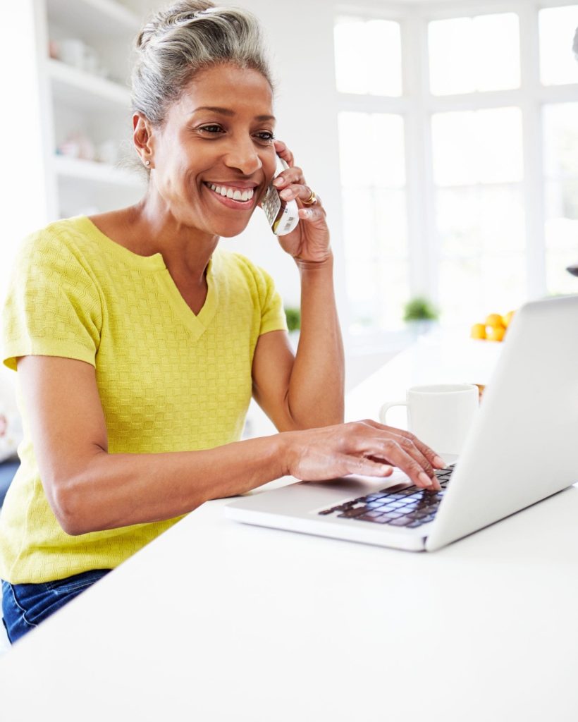 Woman,Using,Laptop,And,Talking,On,Phone,In,Kitchen,At